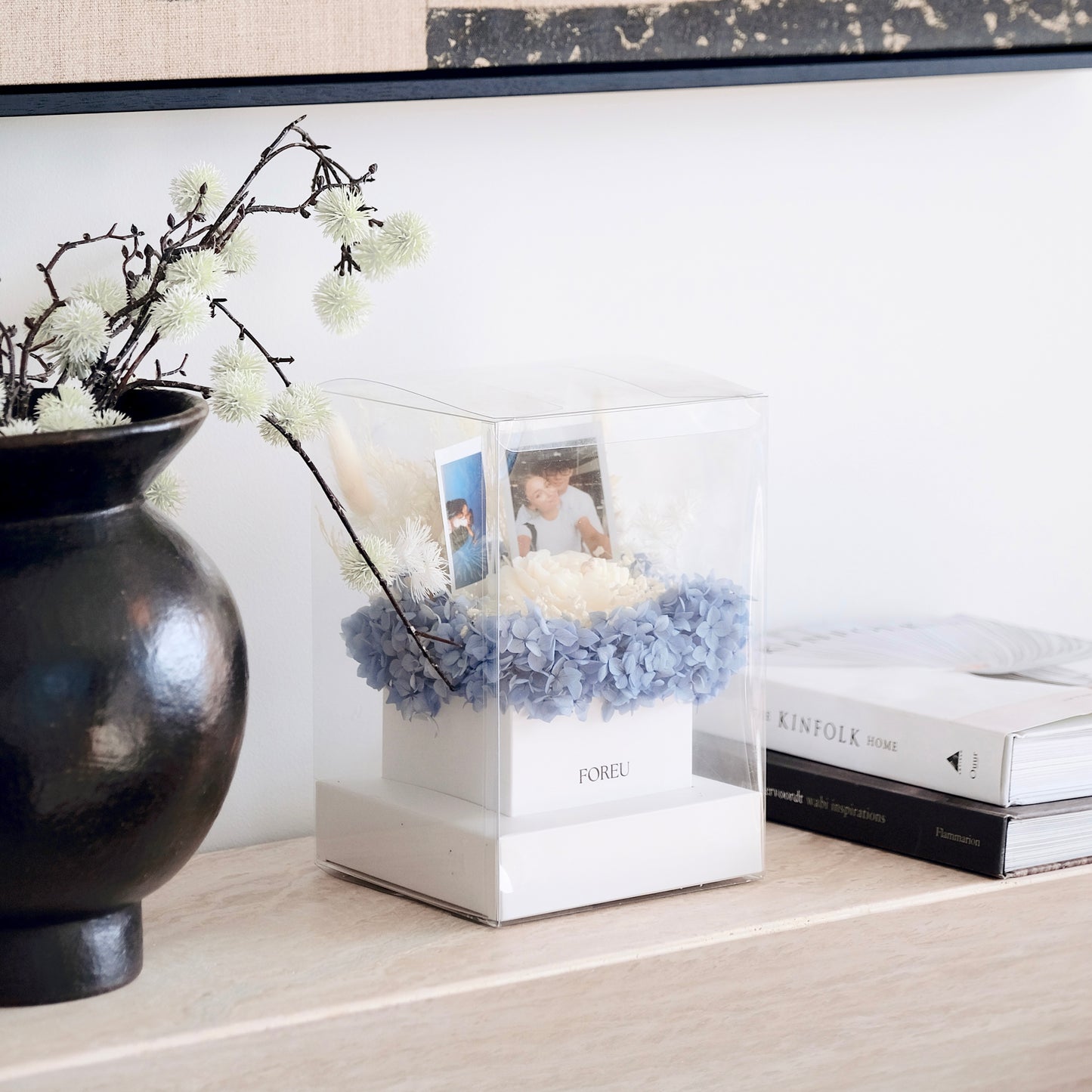 Decorative box with preserved flowers and peony candle and custom polaroid photos on a surface with a vase and books in the background