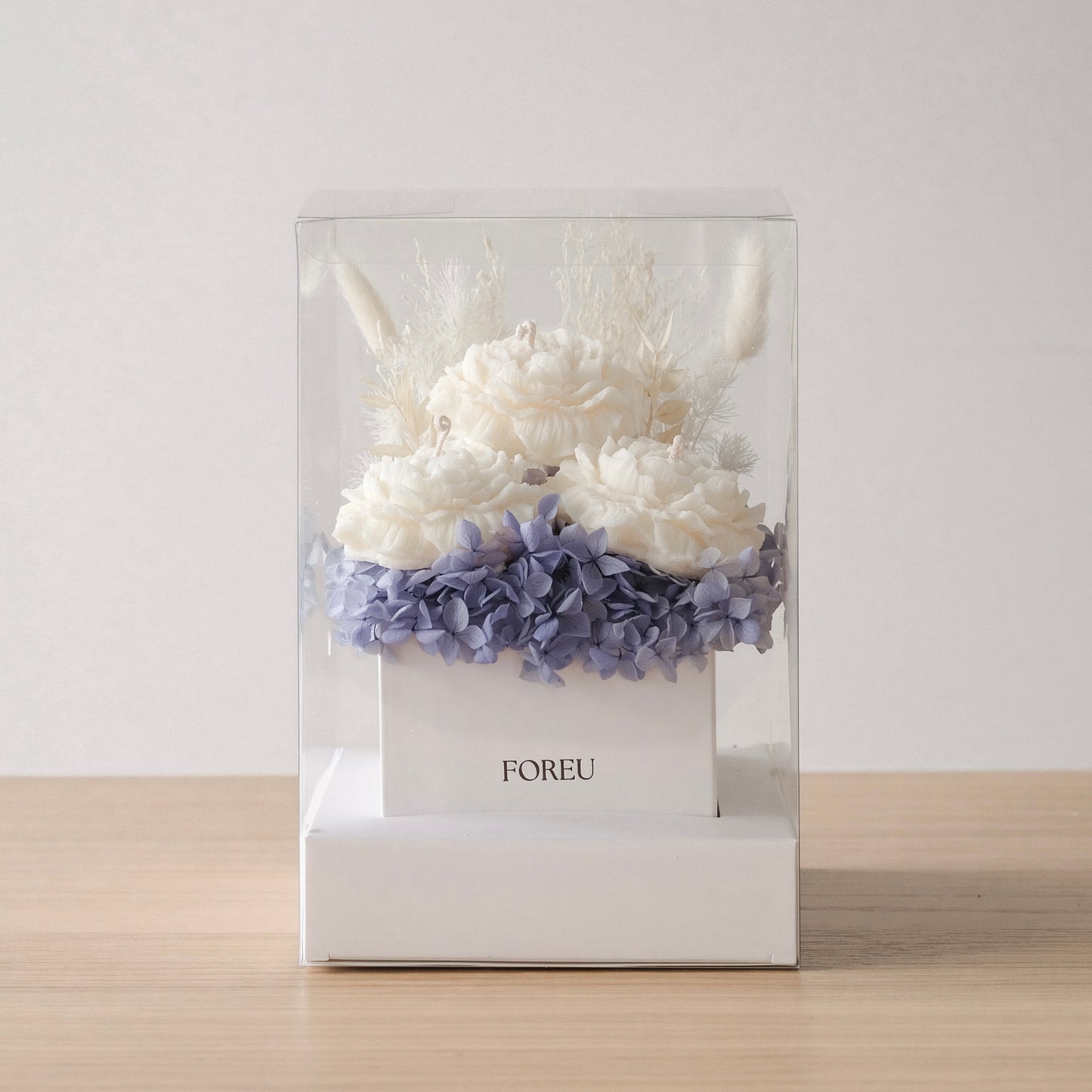 A dried flower and candle box arrangement from FOREU in a Midnight Meadow color scheme, presented in a clear box with a white base, containing white and blue flowers along with decorative candles.