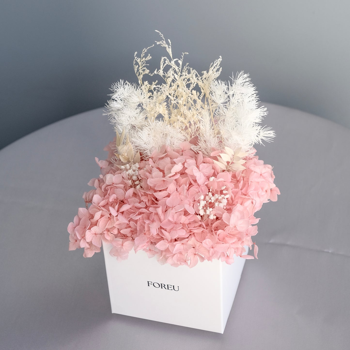 Dried preserved arrangement without outer box plastic packaging in pink colourway. A mix of preserved hydrangeas, bunnies tail, babies breath, ming fern.