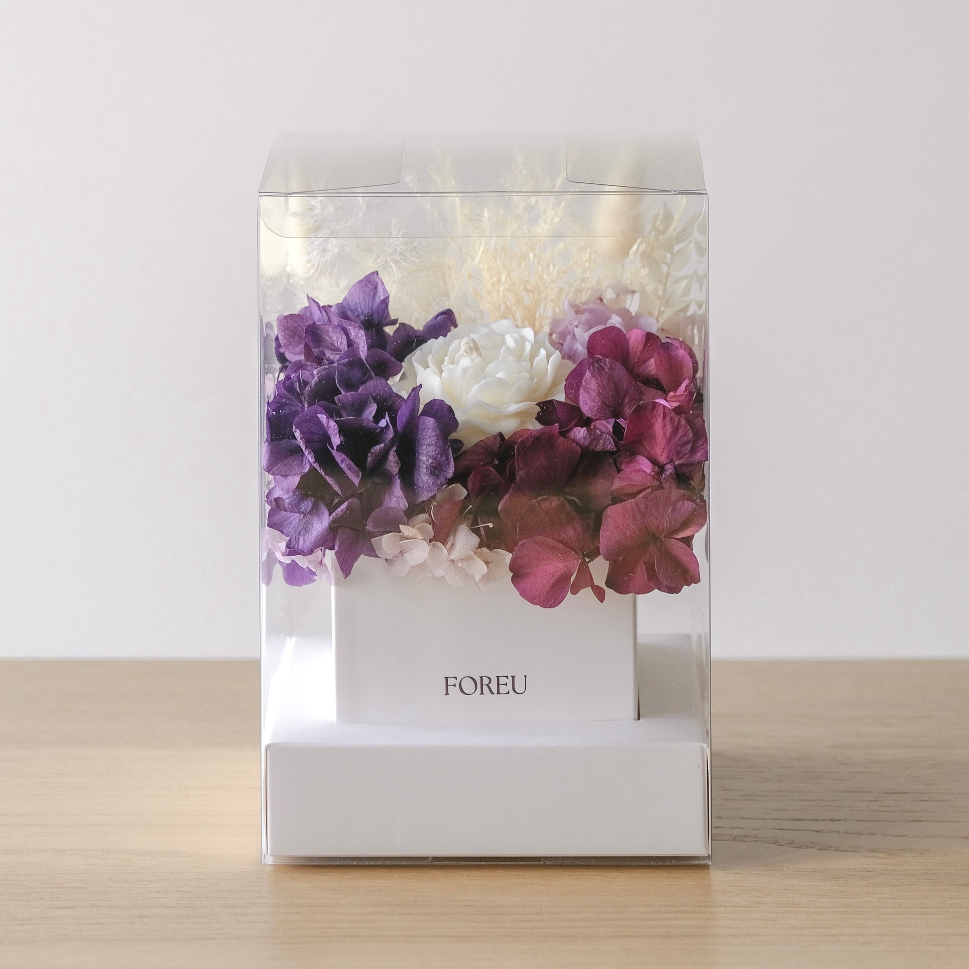 A clear acrylic display box containing a white floral arrangement with purple and burgundy hydrangea-like blooms, set on a light wood surface against a plain light background. The box is branded FOREU on the front.