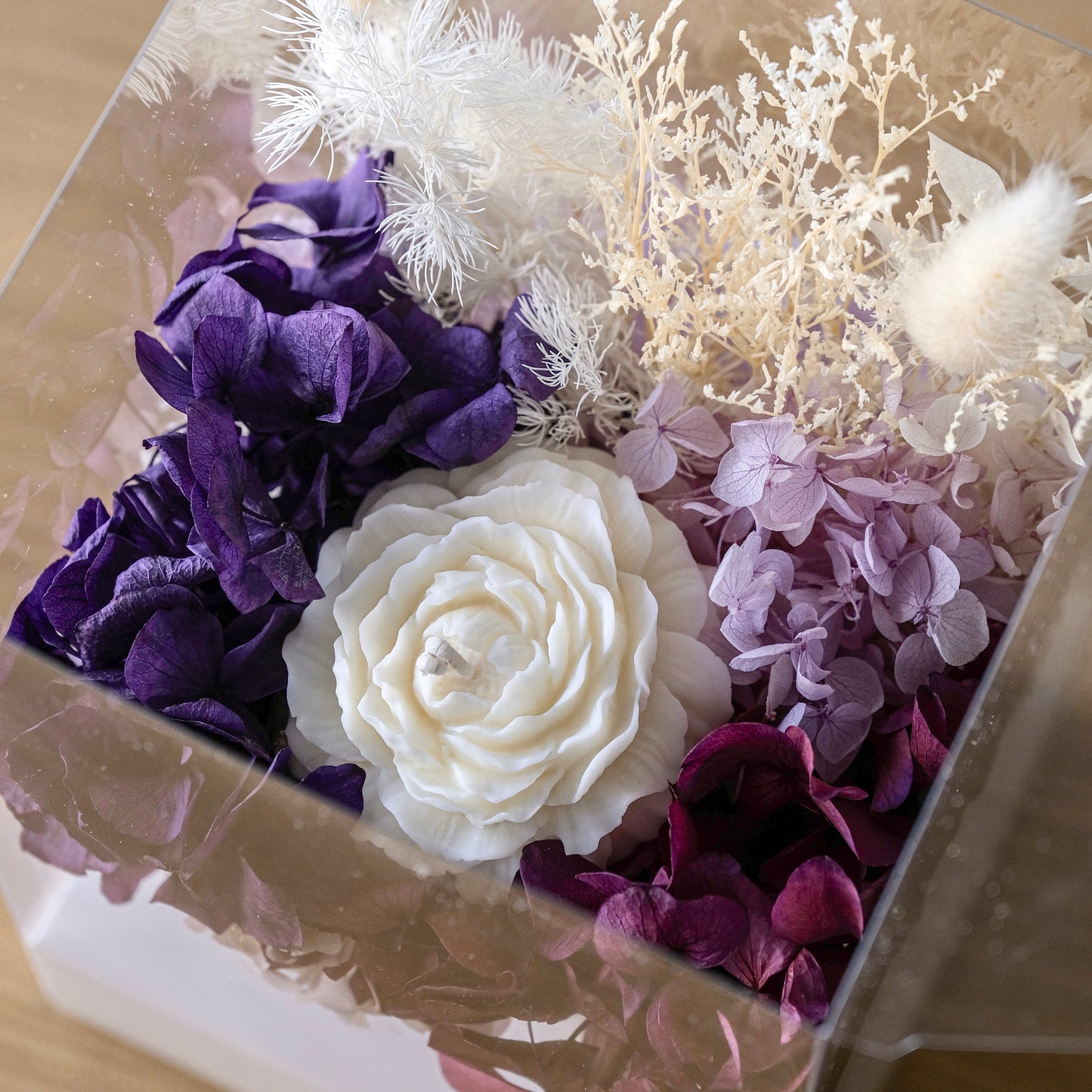 A preserved floral arrangement in a clear square box, featuring deep purple and lavender hydrangeas, a large white rose, and white feathery decorative stems.