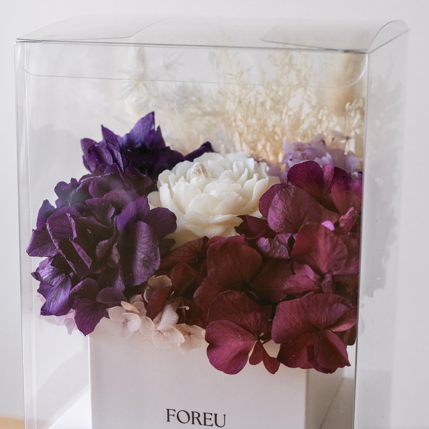 A floral arrangement in a clear square display box with a white base. The bouquet features large purple and deep burgundy hydrangea-like blooms, a central white flower, and pale cream filler stems.