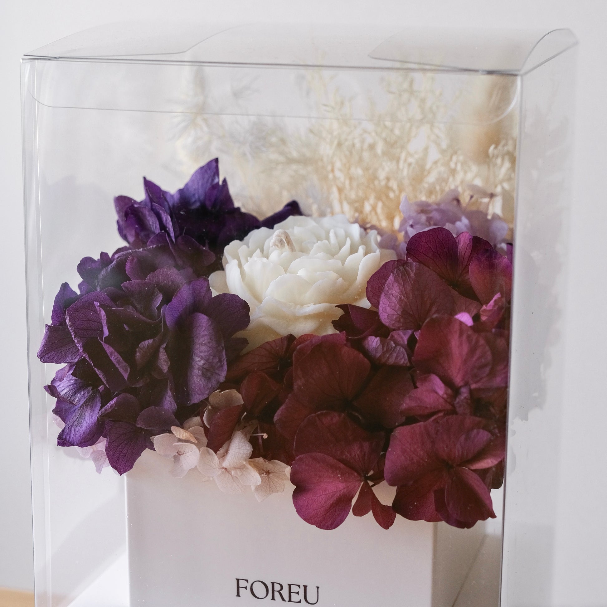 A floral arrangement in a clear square display box with a white base. The bouquet features large purple and deep burgundy hydrangea-like blooms, a central white flower, and pale cream filler stems.