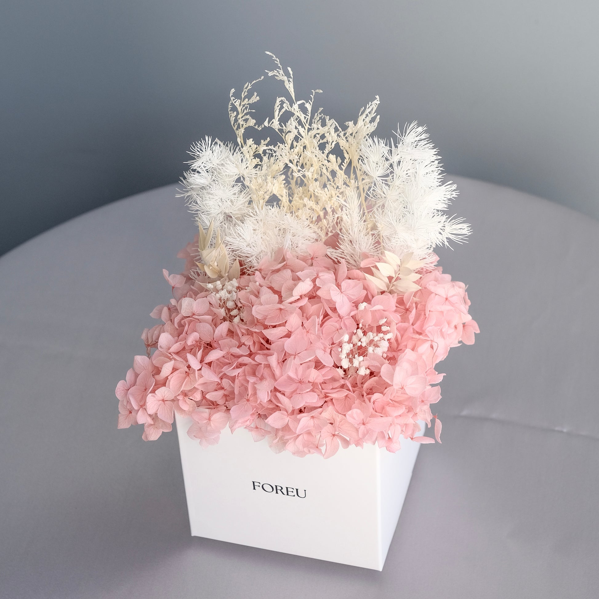 Dried preserved arrangement without outer box plastic packaging in pink colourway. A mix of preserved hydrangeas, bunnies tail, babies breath, ming fern.
